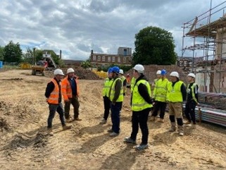 A group of students in yellow hi viz vests facing two construction workers in orange hi viz vests on a construction site